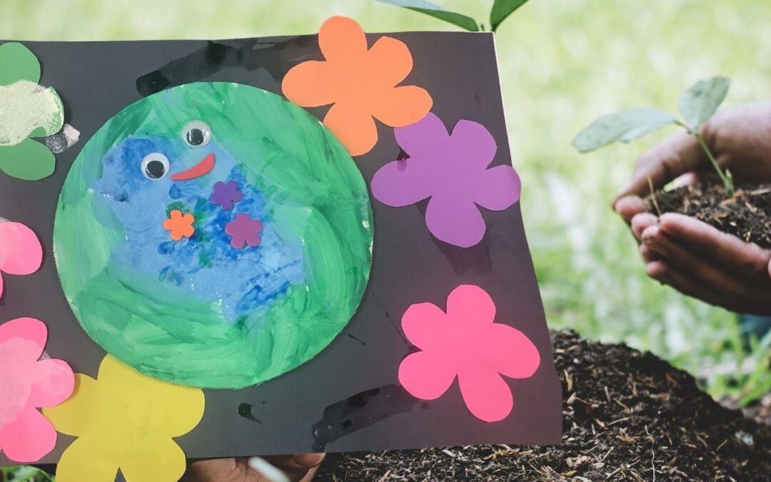 Preschool Earth Day art project featuring a painted Earth with googly eyes and paper flowers, held in front of hands planting a young seedling in soil.